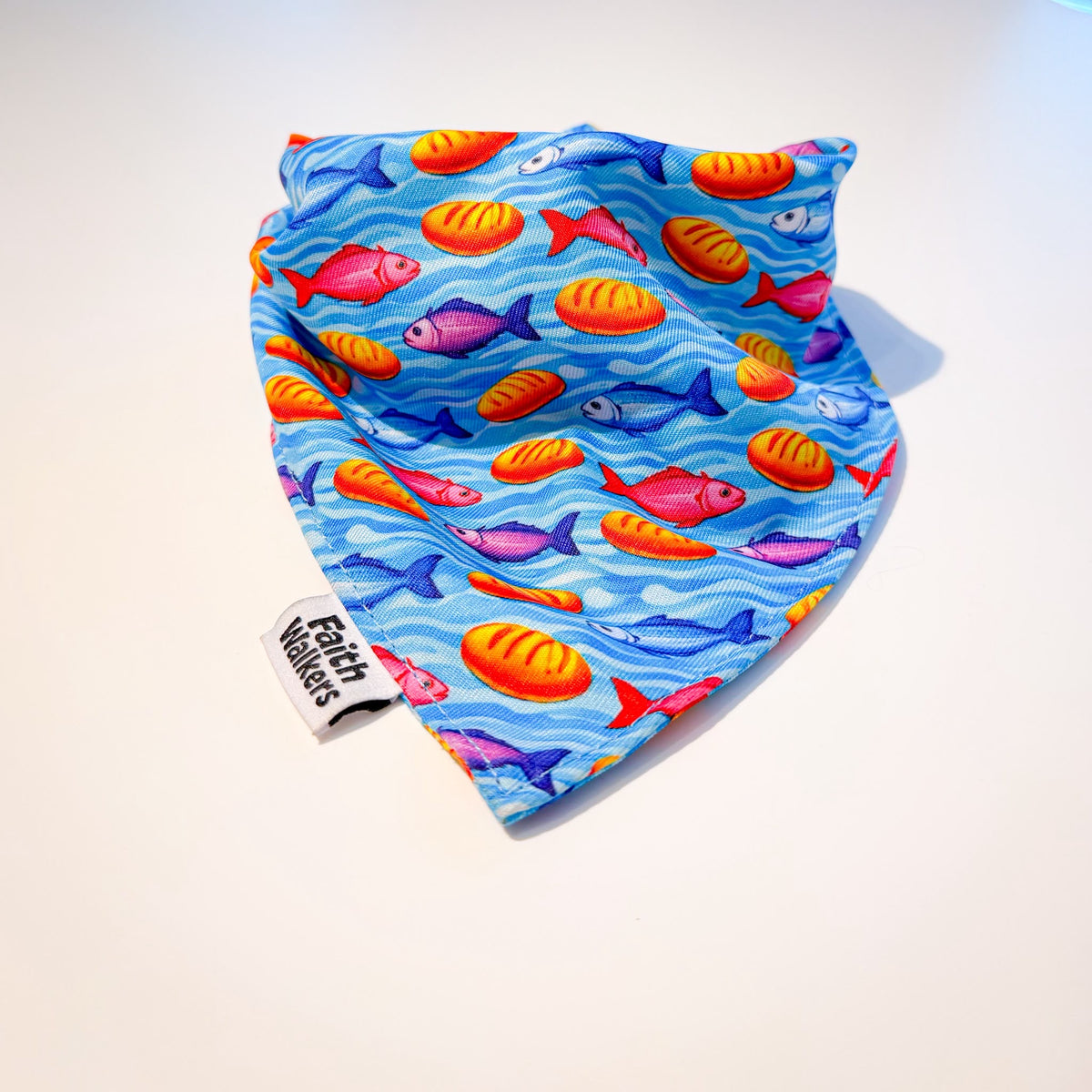 Blue Loaves and Fishes dog bandana with colorful fish, orange bread loaves and a Faith Walkers label on a white background.